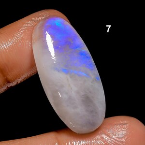 May include: An oval-shaped, polished gemstone with a pearly white base and iridescent blue and purple flashes. The gemstone is held between two fingers against a black background. The number "7" is visible on the upper right.
