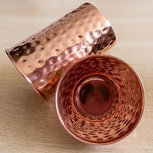 May include: Two cylindrical copper tumblers featuring a distinct hammered texture across their exterior surfaces. The metallic finish reflects light, showcasing the warm reddish-brown tones of the copper material in a bright, clear display.