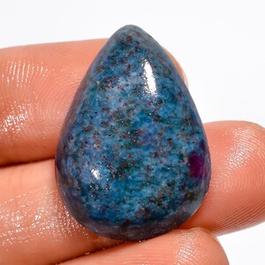 May include: A teardrop-shaped gemstone with a polished surface. The stone features a mottled pattern of blue, teal, and brown hues, with a small, deep red inclusion near the base. The gem is held between fingers.