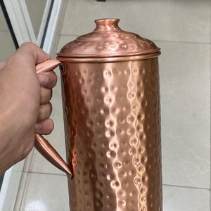 May include: A copper-colored pitcher with a lid and handle. The pitcher has a hammered texture and a cylindrical shape. The handle is attached to the side of the pitcher. The lid sits on top of the pitcher. The pitcher is held by a person.