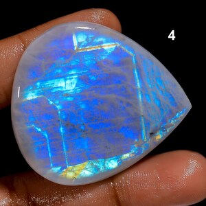 May include: A pear-shaped, polished gemstone with a milky white base and vibrant blue and yellow internal patterns. The stone has a smooth, reflective surface, with the number "4" visible in the upper right corner. The gemstone is held in a hand.