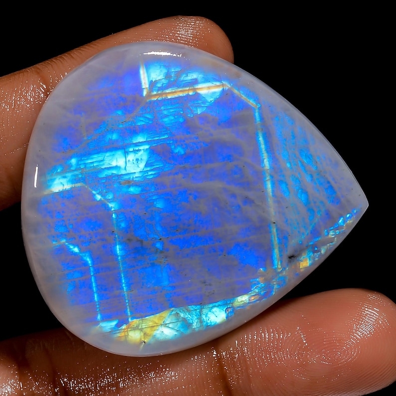 May include: A pear-shaped, polished moonstone gemstone with a milky white base and iridescent blue and yellow flashes. The stone is held against a dark background, showcasing its natural beauty and color variations.