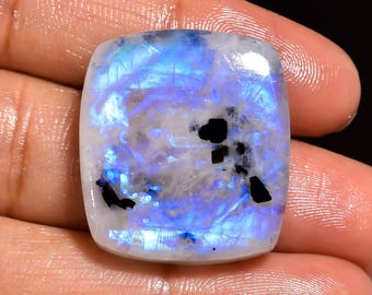 May include: A white moonstone cabochon with blue and iridescent flashes. The stone has black inclusions and is cut in a square shape.