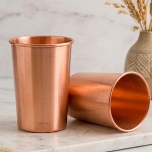 Puede incluir: Dos vasos cilíndricos fabricados en cobre pulido con un acabado metálico. Un vaso está de pie mientras que el otro está tumbado de lado. El vaso que está de pie presenta el pequeño texto grabado PURE COPPER cerca de la base.