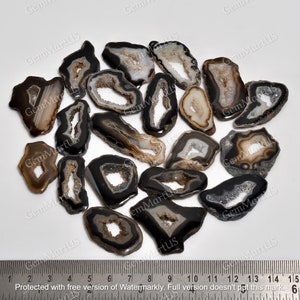 May include: A collection of 17 agate slices, each with a unique shape and size. The agate is black with white and grey banding and has a natural, raw edge. The agate slices are arranged on a white surface with a ruler for scale.