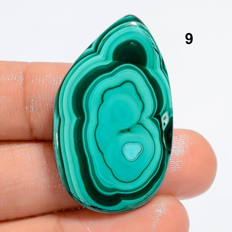 May include: A green malachite cabochon with a swirling pattern. The stone is a teardrop shape and has a smooth, polished surface.