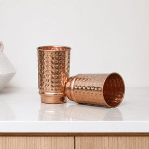 May include: Two copper-colored metal tumblers with a hammered texture. One tumbler stands upright, while the other lies on its side. The tumblers have a cylindrical shape with a slightly flared rim and base. The background is a neutral white.