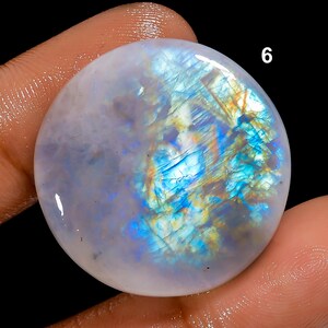 May include: A round, polished moonstone gemstone with a milky white base and iridescent blue, yellow, and orange flashes. The number "6" is visible in white on the upper right side of the stone. The stone is held by a person's fingers.