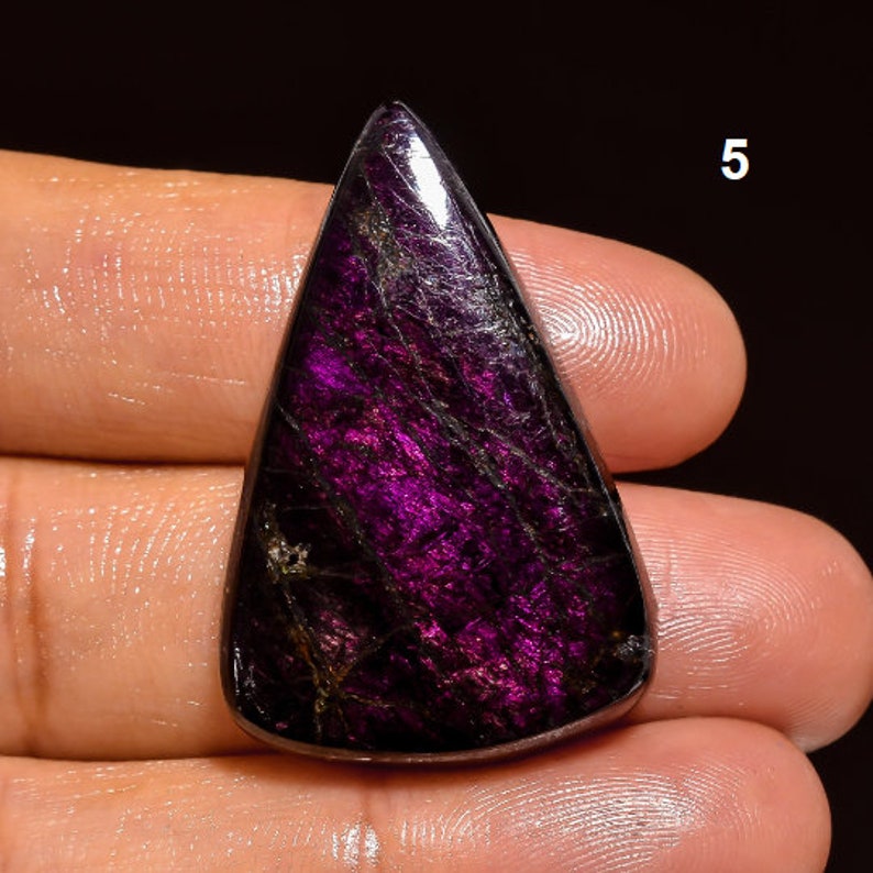 May include: A dark purple and black gemstone cabochon with a triangular shape. The gemstone has a shiny, polished surface and a unique, striated pattern.