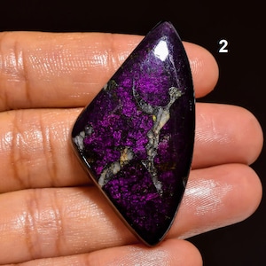May include: A purple and black cabochon gemstone with a triangular shape and a shiny, iridescent finish. The stone has a unique pattern of white and silver veins running through it.
