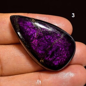 May include: A teardrop-shaped purple and black gemstone with gold flecks. The gemstone is set in a cabochon cut.