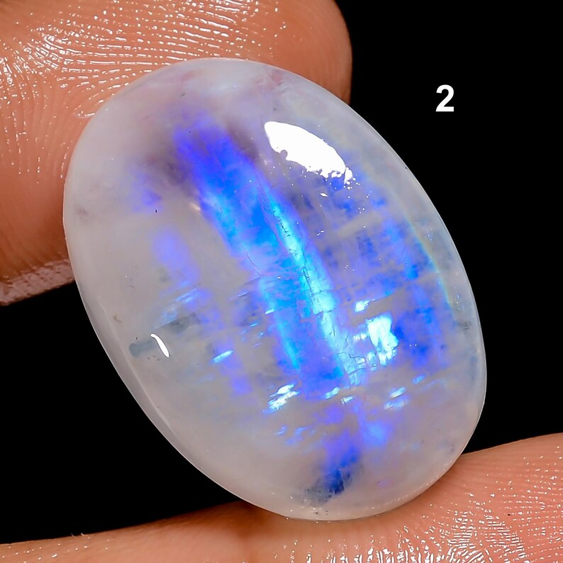K&ouml;nnte beinhalten: Ein oval geschliffener Mondstein mit milchig-wei&szlig;er Basis und irisierenden blauen Reflexen. Die Edelstein wird zwischen Fingern gehalten, mit der Nummer "2" im Hintergrund. Die Oberfl&auml;che des Steins reflektiert das Licht und zeigt seine nat&uuml;rliche Sch&ouml;nheit.