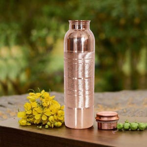 Könnte beinhalten: Eine Kupferwasserflasche mit passendem Deckel. Die Flasche hat eine zylindrische Form mit horizontalen geätzten Linien. Der Deckel ist rund und liegt neben der Flasche. Gelbe Blumen und grüne Beeren befinden sich im Vordergrund.