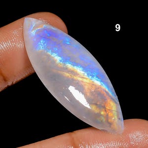 May include: A polished, elongated gemstone with a pearlescent white base and iridescent rainbow flashes of blue, yellow, and orange. The gemstone is held between fingers against a black background, with the number "9" visible.