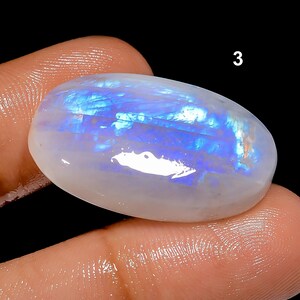 K&ouml;nnte beinhalten: Ein polierter, oval geformter Edelstein mit einer perlmuttwei&szlig;en Basis und irisierenden blauen und wei&szlig;en Innenmustern. Der Edelstein wird zwischen Fingern vor einem schwarzen Hintergrund gehalten. Die Zahl "3" ist oben rechts sichtbar.