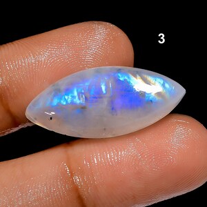 May include: A polished, oval-shaped moonstone gemstone with a pearlescent white base and iridescent blue and purple flashes. The gemstone is held between two fingers against a black background. The number "3" is visible in the upper right corner.