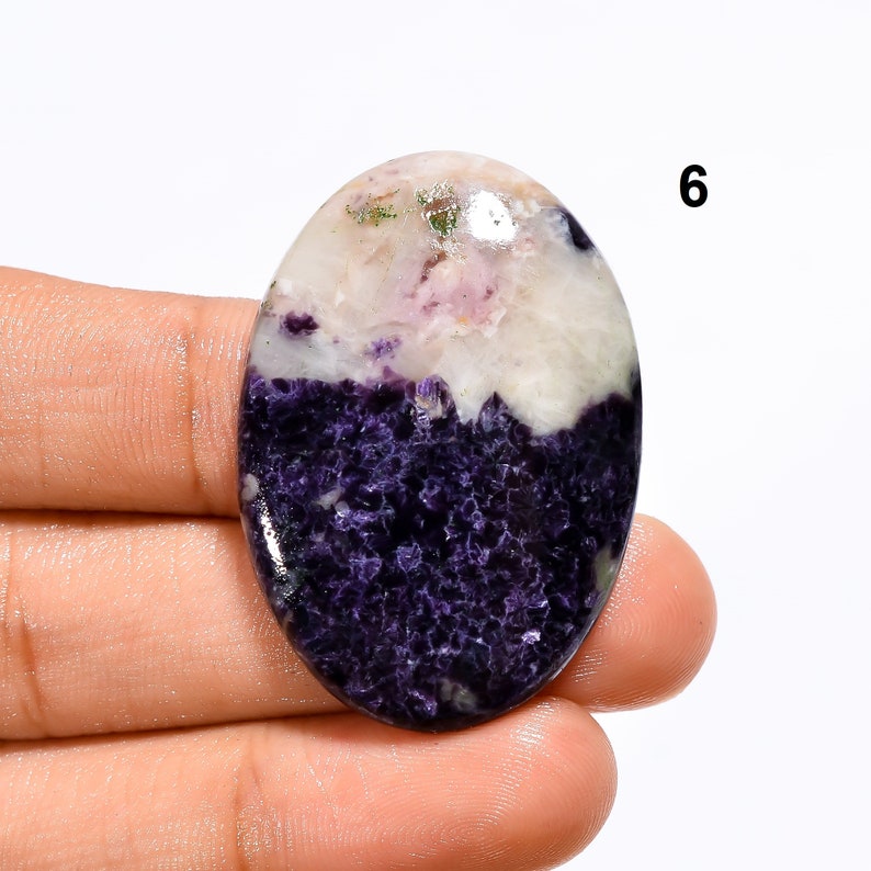 May include: Oval shaped purple and white gemstone cabochon with a rough, textured surface. The stone has a white top with purple and pink swirls and a purple bottom with a rough, textured surface.
