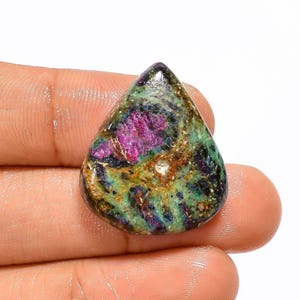 May include: A teardrop-shaped cabochon gemstone with a green, brown, and pink colour pattern. The gemstone has a rough, natural finish.