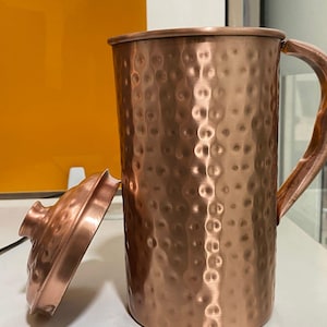 May include: A copper-colored pitcher with a hammered texture and a matching lid. The pitcher has a wooden handle and is sitting on a white surface. The lid is slightly ajar, revealing the interior. The pitcher is approximately 8 inches tall.