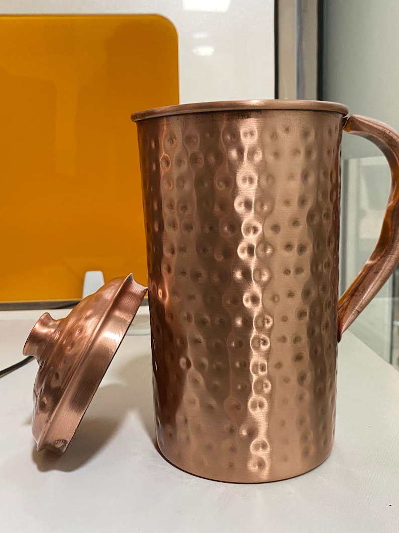 May include: A copper-colored pitcher with a hammered texture and a matching lid. The pitcher has a wooden handle and is sitting on a white surface. The lid is slightly ajar, revealing the interior. The pitcher is approximately 8 inches tall.