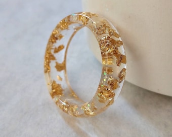 May include: A clear resin ring with gold flakes embedded inside. The ring is thick and has a rounded shape.