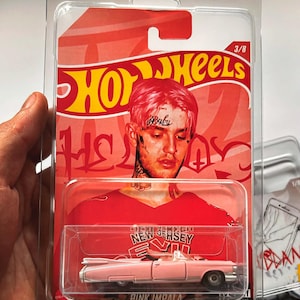 May include: A Hot Wheels die-cast toy car in a clear plastic package. The packaging features a portrait of a person with pink hair and the text "Hot Wheels". The car is a pink convertible labeled "Pink Impala".
