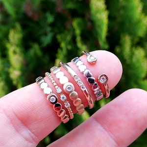May include: A stack of six gold and silver rings with small diamonds on a finger. The rings are all different styles, including a ring with a row of diamonds, a ring with a row of small circles, and a ring with a row of hearts.