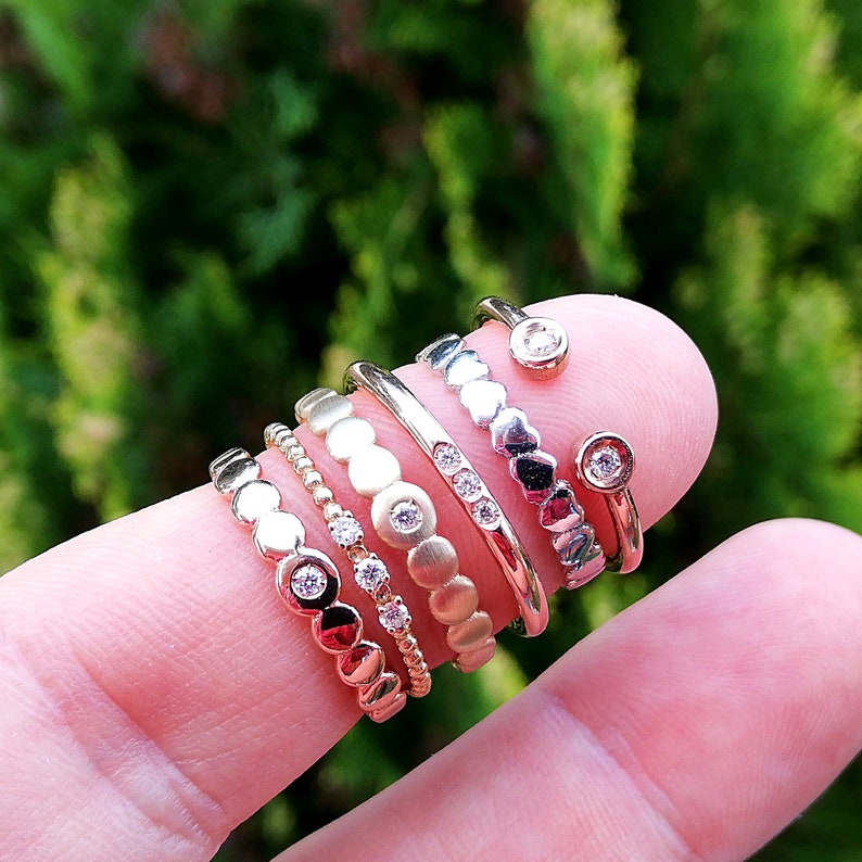 May include: A stack of six gold and silver rings with small diamonds on a finger. The rings are all different styles, including a ring with a row of diamonds, a ring with a row of small circles, and a ring with a row of hearts.