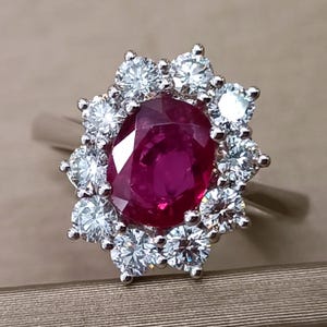 May include: A ring with a central oval ruby, encircled by a halo of smaller, brilliant-cut diamonds. The ruby is a rich red hue, and the diamonds provide a sparkling contrast. The ring is set in a silver band.