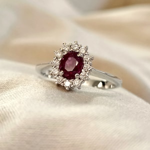 Può includere: Un anello d'argento con un grande rubino ovale circondato da un'aureola di diamanti più piccoli.