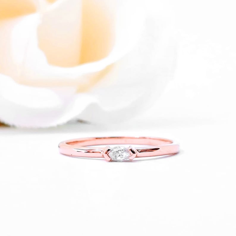 May include: A delicate rose gold band ring with a single, small, clear marquise-shaped diamond set in the center.
