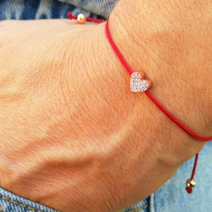 May include: A red string bracelet with a small heart-shaped charm set with clear cubic zirconia stones. The charm is attached to the string with a gold-toned metal clasp.