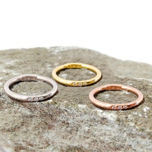 May include: Three simple wedding bands with three small diamonds set in a row on each band. The bands are shown on a gray rock. The bands are in silver, gold, and rose gold.