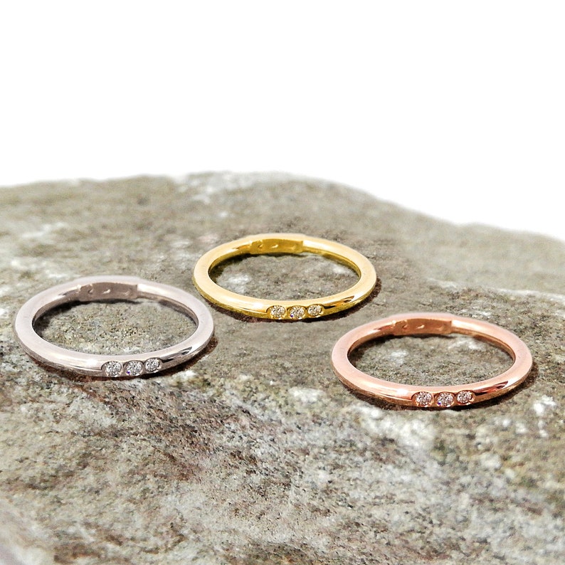 May include: Three simple wedding bands with three small diamonds set in a row on each band. The bands are shown on a gray rock. The bands are in silver, gold, and rose gold.