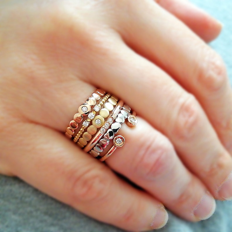 May include: A stack of five gold, silver, and rose gold rings with small round stones set in each band. The rings are worn on a person's finger.