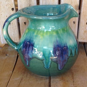 May include: A handcrafted ceramic pitcher with a large handle and a pouring spout. The pitcher features a glossy glaze in shades of teal, green, and blue, with darker purple drips. The pitcher is a decorative item.
