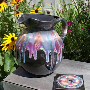 May include: A black ceramic pitcher with a handle and a spout, featuring a colorful, dripped glaze in shades of purple, blue, and orange. A matching coaster with a similar design sits nearby. The pitcher is displayed outdoors.