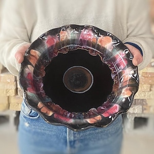 May include: A black ceramic bowl with a scalloped rim. The bowl has a colorful glaze with shades of pink, orange, and green. The inside of the bowl is black.