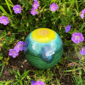 May include: A green, blue, and yellow ceramic sphere with a swirling pattern. The sphere is sitting on a bed of green grass and purple flowers.