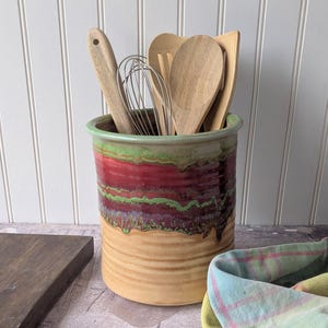 May include: A ceramic utensil holder with a green rim, red and purple bands, and a tan base. It contains wooden spoons, a whisk, and spatulas. The holder sits on a countertop with a wooden cutting board and a plaid cloth nearby.