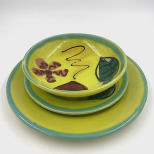 May include: A ceramic dinnerware set consisting of a large plate, a smaller plate, and a bowl, all featuring a bright yellow glaze with a light blue rim. The pieces are decorated with hand-painted brown floral motifs and green leaf designs.