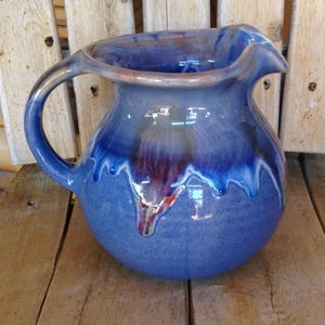 May include: A blue ceramic pitcher with a rounded body and a curved handle. The pitcher has a glossy finish with darker blue and purple drips. The pitcher is sitting on a wooden surface.