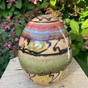 May include: A ceramic egg with a colorful, abstract design. The egg is painted with shades of pink, green, blue, and black. It has a glossy finish.