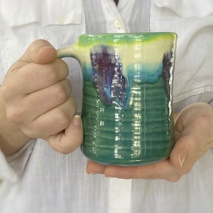 May include: Handmade ceramic mug with a unique glaze. The mug features a green base with blue, purple, and cream-colored accents. The mug has a textured, ridged design and a sturdy handle. Perfect for coffee, tea, or any beverage.
