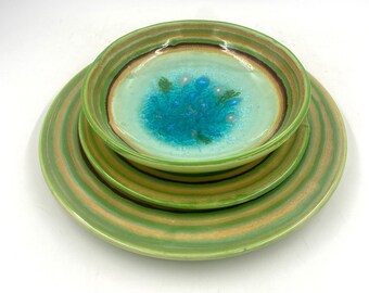 May include: A stack of three ceramic dishes. The largest plate is green with horizontal stripes of yellow and brown. The medium bowl has a light green center with a blue and teal design. The smallest bowl is stacked on top.