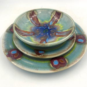 May include: A set of three ceramic dishes: a large plate, a medium plate, and a bowl. The dishes have a teal base color with a colorful, abstract design in shades of blue, purple, and yellow. The set is suitable for serving food.