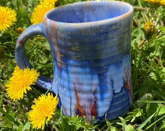 May include: A blue and white ceramic mug with a colorful glaze. The mug has a handle and is sitting on a bed of green grass and yellow dandelions.