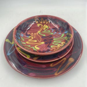 May include: A set of two stacked ceramic plates with a glossy red base glaze. The surface features abstract, hand-painted squiggles and elongated shapes in vibrant yellow, lime green, light blue, and deep purple, creating a colorful pattern.