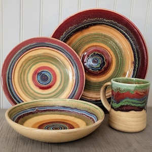May include: A ceramic dinnerware set featuring a large plate, a smaller plate, a bowl, and a mug. The pieces have a concentric circle design in shades of red, green, blue, and yellow. The mug has a tan base.