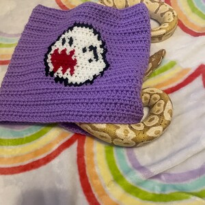 May include: A purple crocheted hat with a cartoon ghost design in white, red, and black. A tan and brown snake is partially visible, coiled around the hat. The background is a soft blanket with a rainbow design.